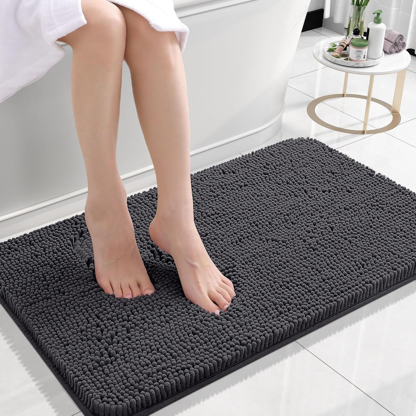 Dark gray absorbent chenille bath mat on white tile in a bathroom beside a vanity
