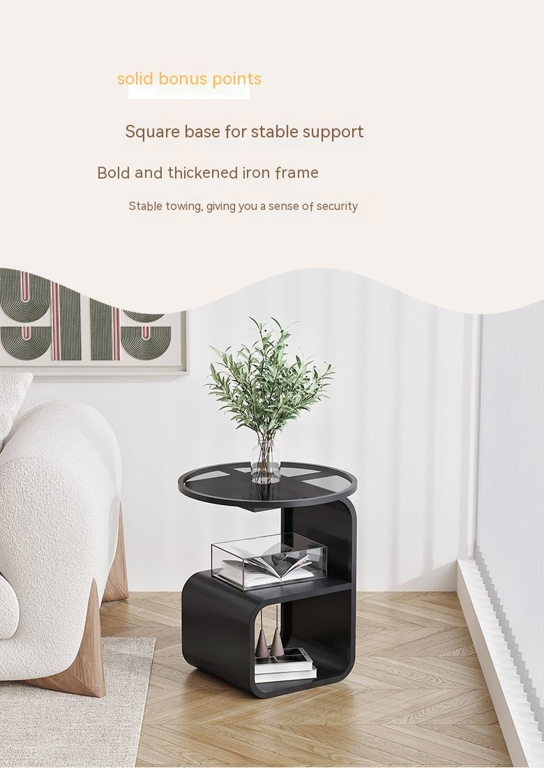 Round glass sofa side table holding a drink, lamp, and remote beside a living room sofa
